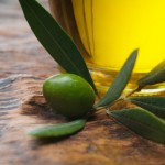 A close up shot of an olive next to a glass bottle of olive oil.