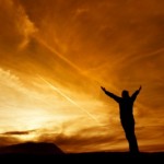 A man standing looking at a sunset with his arms outstretched.