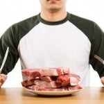 A man sitting down in front of a plate full of raw steak with a knife and fork.