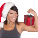 A fit girl in a Christmas hat flexing her arm whilst balancing a present.