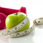 A green apple wrapped in a tape measure next to a pair of pink dumbbells.