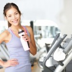 A woman standing on a cardio machine with a bottle of water smiling.