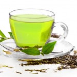 A g;ass cup of green tea surrounded by green tea leaves.